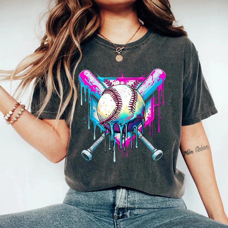 May include: Dark gray t-shirt featuring a baseball design. The graphic includes a baseball with dripping paint, crossed bats, and a shield shape, in shades of blue, pink, and white. The shirt is a casual style.