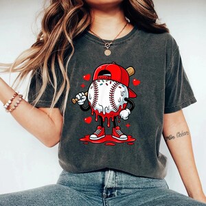 May include: Dark gray t-shirt featuring a baseball character with a red cap and sneakers, holding a bat. The baseball has a white and red design with dripping red accents and is surrounded by red hearts.