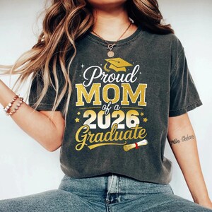 May include: Dark gray t-shirt with the text "Proud MOM of a 2026 Graduate" in gold and white. The design includes a graduation cap and scroll. The shirt is a comfortable, casual style.