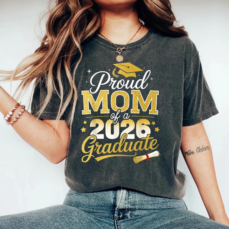 May include: Dark gray t-shirt with the text "Proud MOM of a 2026 Graduate" in gold and white. The design includes a graduation cap and scroll. The shirt is a comfortable, casual style.