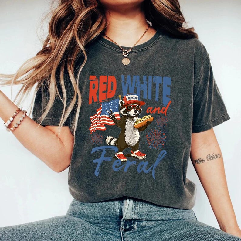 Puede incluir: Camiseta gris oscuro con un gr&aacute;fico que presenta un mapache con sombrero y sosteniendo un perrito caliente, con el texto "Red White and Feral". El mapache est&aacute; frente a una bandera estadounidense y fuegos artificiales.
