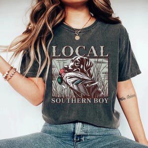 Pu&ograve; includere: T-shirt grigio scuro con una grafica di un cane da caccia con un'anatra in bocca, sullo sfondo di canne. Le parole "LOCAL" e "SOUTHERN BOY" sono stampate sopra e sotto l'immagine in un carattere serif.