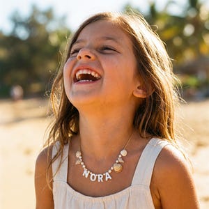 Puede incluir: Un collar dorado con el nombre "NORA" en letras blancas, con detalles de conchas marinas y perlas. La niña lleva un vestido color crema y está sonriendo. El fondo es una escena de playa.