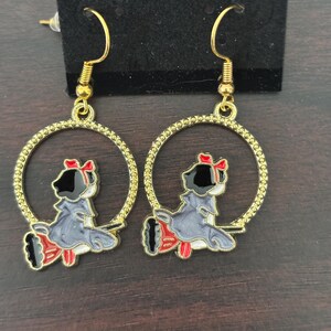 May include: A pair of gold-colored hoop earrings. Each earring features a cartoon figure in a gray dress with a red bow and accents. The figure is inside a gold-colored hoop with a textured edge. The earrings are displayed on a black card.