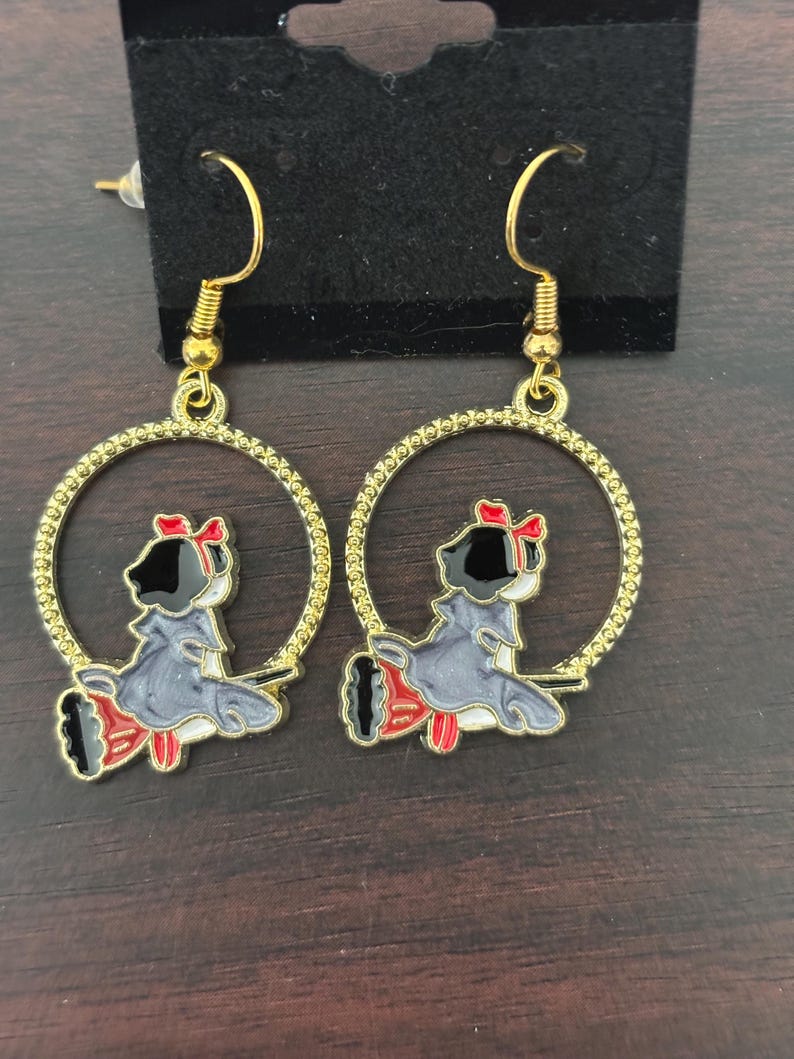 May include: A pair of gold-colored hoop earrings. Each earring features a cartoon figure in a gray dress with a red bow and accents. The figure is inside a gold-colored hoop with a textured edge. The earrings are displayed on a black card.