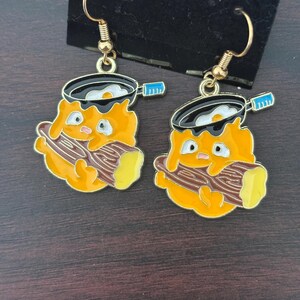 May include: A pair of gold-colored dangle earrings. Each earring features a cartoon character with an orange body, holding a log, and a frying pan with a fried egg. The pan has a black rim and a blue handle. The earrings are on a black display card.
