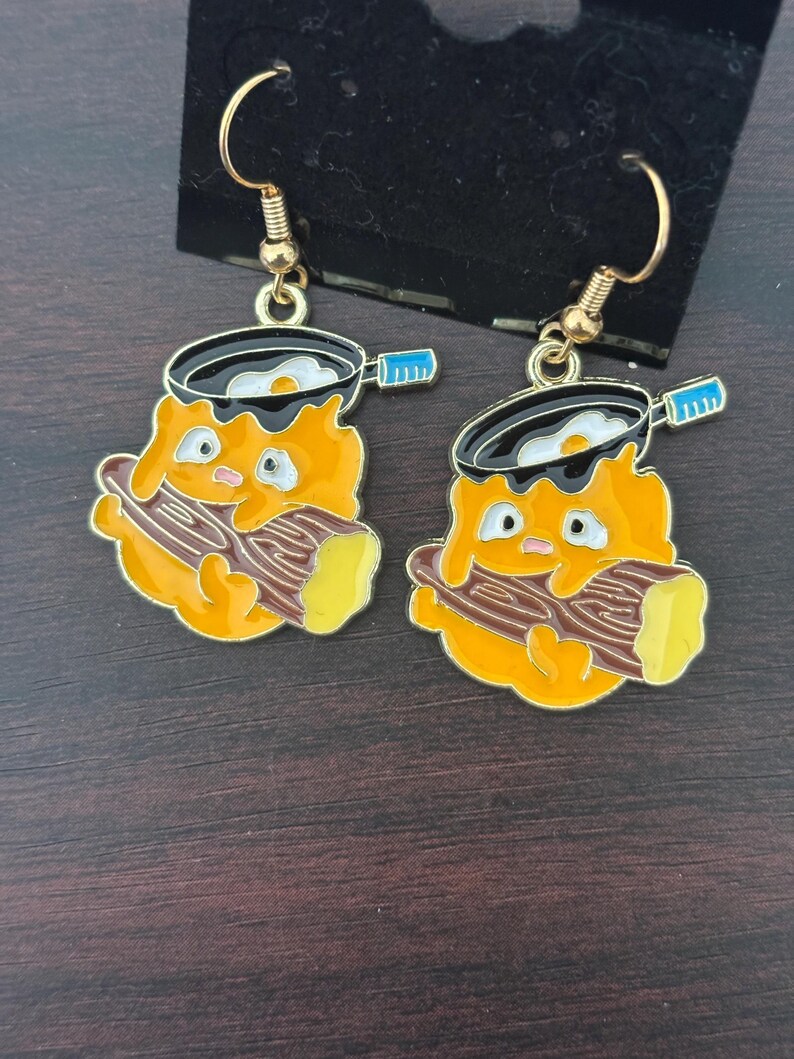 May include: A pair of gold-colored dangle earrings. Each earring features a cartoon character with an orange body, holding a log, and a frying pan with a fried egg. The pan has a black rim and a blue handle. The earrings are on a black display card.