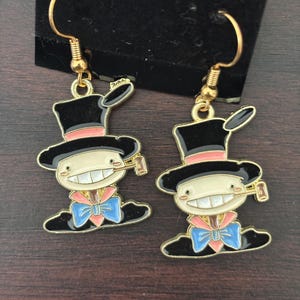 May include: A pair of gold-tone dangle earrings. Each earring features a cartoon character with a black top hat, a pink band, and a blue bow tie. The character has a wide smile and is holding a small object.