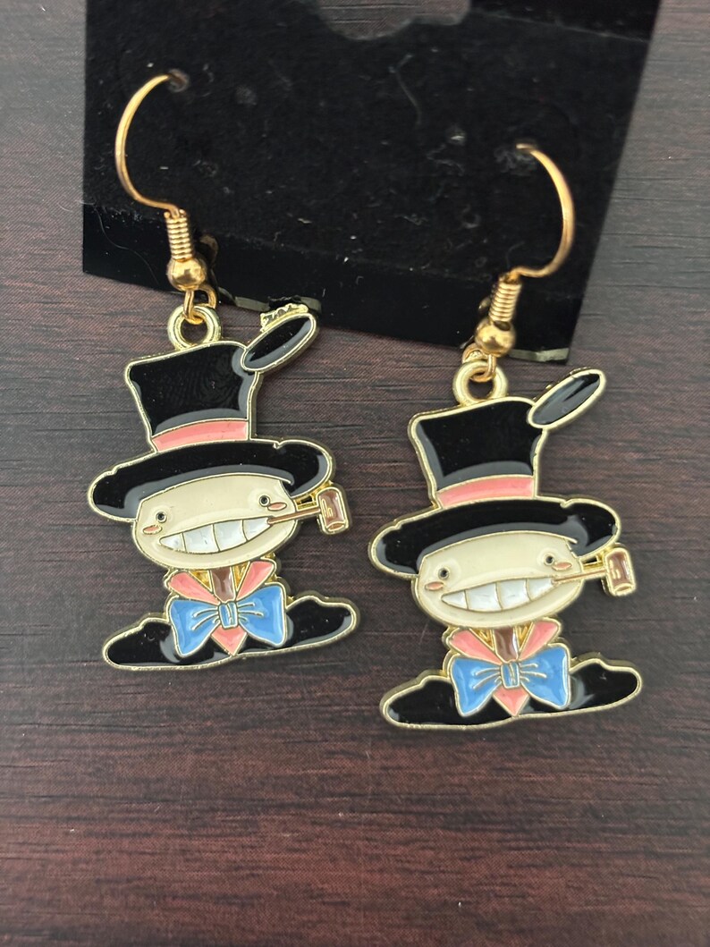 May include: A pair of gold-tone dangle earrings. Each earring features a cartoon character with a black top hat, a pink band, and a blue bow tie. The character has a wide smile and is holding a small object.