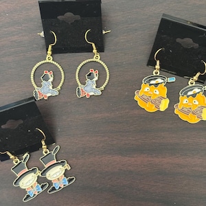 May include: Gold-tone dangle earrings featuring enamel designs. The earrings showcase three different designs: a girl in a blue dress, a chef with a toothbrush, and a man in a top hat. Each earring is attached to a black display card.