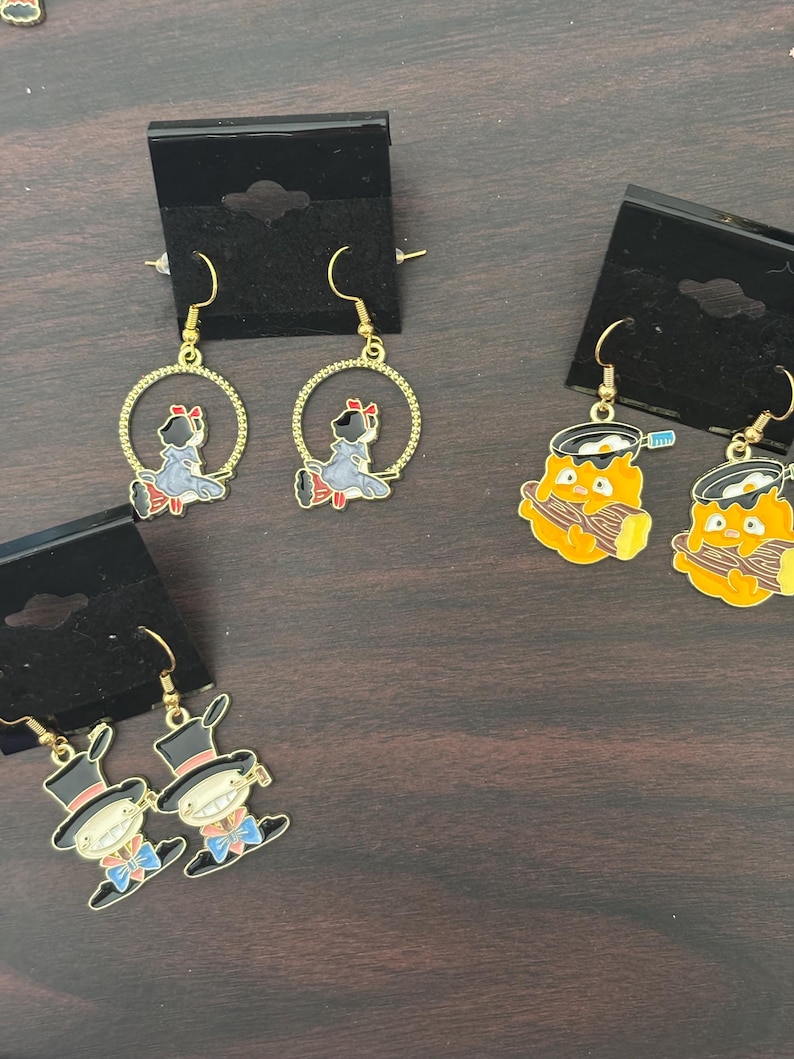 May include: Gold-tone dangle earrings featuring enamel designs. The earrings showcase three different designs: a girl in a blue dress, a chef with a toothbrush, and a man in a top hat. Each earring is attached to a black display card.