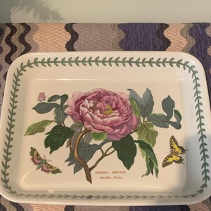 May include: A white ceramic baking dish with a green border and a floral design featuring a pink peony flower and a yellow butterfly. The dish has the text "PEONIA MOUTAN Shrubby Peony" printed on the bottom.