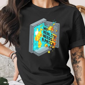 May include: Black t-shirt with the text "MY REALITY NEEDS A PATCH" and a graphic of an open safe filled with colorful 3D blocks. The design features blue, yellow, and orange cubes, creating a pixelated effect.