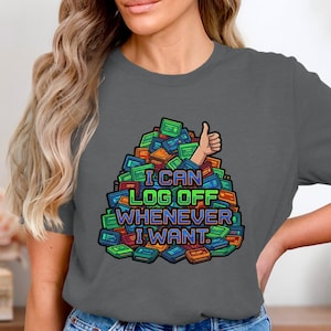 May include: Dark grey t-shirt with a colourful graphic. The graphic shows a pile of retro video game cartridges with the text "I CAN LOG OFF WHENEVER I WANT" and a thumbs-up hand gesture.