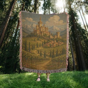 May include: A tapestry throw blanket depicting a castle on a hill, vineyards, and mountains. The blanket has a fringed edge with pink and tan threads. The background is a forest with tall trees and sunlight. The blanket is made of woven fabric.