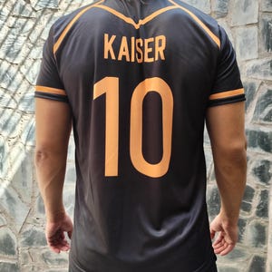 以下が含まれることがあります： オレンジ色のアクセントが付いた黒色のスポーツジャージ。名前「KAISER」と番号「10」がオレンジ色で表示されています。袖と首周りにはオレンジ色のトリムが付いています。光沢のあるアスレチック素材で作られています。