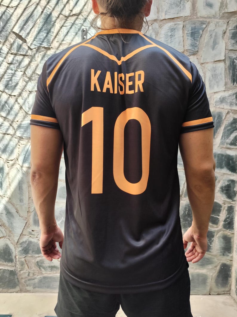 以下が含まれることがあります： オレンジ色のアクセントが付いた黒色のスポーツジャージ。名前「KAISER」と番号「10」がオレンジ色で表示されています。袖と首周りにはオレンジ色のトリムが付いています。光沢のあるアスレチック素材で作られています。