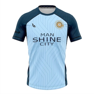 May include: Light blue and navy blue soccer jersey with the text "MAN SHINE CITY" and a circular emblem. The jersey has a vertical striped pattern on the front and diamond-patterned shoulders. The sleeves and collar are navy blue.