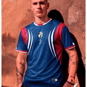 K&ouml;nnte beinhalten: Ein kurz&auml;rmeliges Sporttrikot in Blau, Rot und Wei&szlig; mit einem sich wiederholenden geometrischen Muster. Auf der Brust befindet sich ein goldenes Hahn-Emblem &uuml;ber einer kleinen franz&ouml;sischen Flagge. Der Text umfasst "2026", "LOOK ORIGINALS", "COLECC.MUNDIAL", "THERMOFIT-TECH", "DISTINTO. UNICO" und "PRES: FRANCIA MUNDIAL".