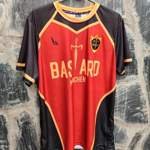 以下が含まれることがあります： 赤と黒のジャージで、オレンジ色のアクセントが施されています。「BASTARD」の文字が大きなオレンジ色の文字でプリントされ、その上に剣のグラフィックがあります。ジャージにはシールドエンブレムと「MUNCHEN」の文字も付いています。