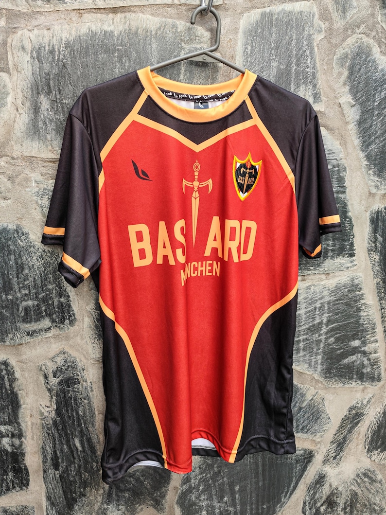 以下が含まれることがあります： 赤と黒のジャージで、オレンジ色のアクセントが施されています。「BASTARD」の文字が大きなオレンジ色の文字でプリントされ、その上に剣のグラフィックがあります。ジャージにはシールドエンブレムと「MUNCHEN」の文字も付いています。