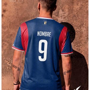 K&ouml;nnte beinhalten: Ein blaues Sporttrikot mit roten Seitenteilen und wei&szlig;en Abschl&uuml;ssen an den &Auml;rmeln. Auf der R&uuml;ckseite befindet sich der wei&szlig;e Schriftzug NOMBRE &uuml;ber der gro&szlig;en wei&szlig;en Nummer 9. Ein dezentes geometrisches Muster bedeckt den blauen Stoff.