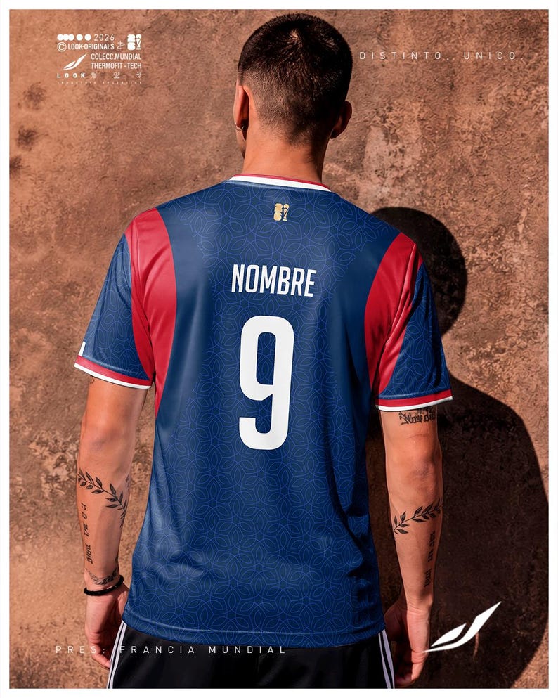 K&ouml;nnte beinhalten: Ein blaues Sporttrikot mit roten Seitenteilen und wei&szlig;en Abschl&uuml;ssen an den &Auml;rmeln. Auf der R&uuml;ckseite befindet sich der wei&szlig;e Schriftzug NOMBRE &uuml;ber der gro&szlig;en wei&szlig;en Nummer 9. Ein dezentes geometrisches Muster bedeckt den blauen Stoff.