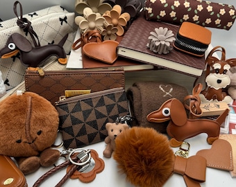 May include: A collection of brown and tan accessories, including wallets, keychains, and a small notebook. The items feature various textures and patterns, such as a dachshund-shaped keychain, a fluffy pom-pom, and a floral-patterned pencil case.