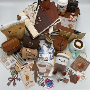 May include: A selection of coffee-themed accessories, such as notebooks, plush toys, keyrings, stickers, and small bags. The items display coffee cups, beans, and related designs in brown, beige, and white. Some items have the text "Coffee & a friend, the perfect blend" and "Life Begins".