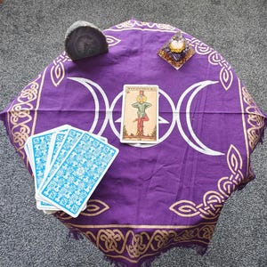 Op de afbeelding: Een paarse tarotdoek met een drievoudig maandesign en gouden rand. Er liggen tarotkaarten en een kristal op de doek. De tekst op de kaart luidt "De Gehangene".