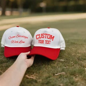 May include: Two white baseball caps with red peaks. One cap has the text "From Saratoga With Love" in red script, and the other says "CUSTOM YOUR TEXT" in red block letters. Both caps have a red button on top.
