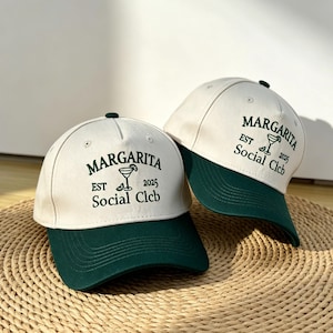 Op de afbeelding: Twee baseballpetten met een crèmekleurige kroon en een donkergroen vizier. De petten hebben de tekst "Margarita Est 2025 Social Clcb" in het groen, samen met een margarita-glasafbeelding. De petten worden gepresenteerd op een geweven oppervlak.