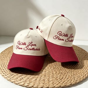 May include: Two baseball caps with cream-colored crowns and dark red brims. The front of each cap features the embroidered text "With Love, From Scottsdale" in a dark red script font. The side of the right cap displays the text "Custom Text".