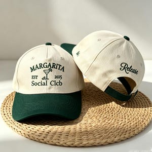 May include: Two baseball caps with a cream-coloured crown and a dark green brim. The front cap features the text "MARGARITA EST 2025 Social Clcb" in dark green. The back cap has the word "Release" embroidered in a script font.