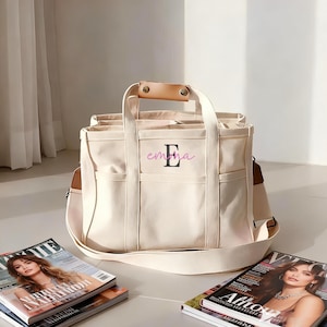 May include: Cream canvas tote bag with multiple pockets and a shoulder strap. The bag has a leather handle and the name "Emma" in pink script with a black initial "E". Magazines are placed near the bag.