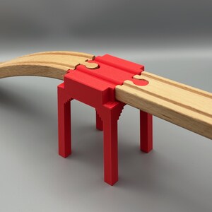 Könnte beinhalten: Eine rote, 3D-gedruckte Brücke trägt eine hölzerne Spielzeugeisenbahnstrecke. Die Brücke hat ein rechteckiges Design mit vier Beinen und einem oberen Abschnitt, der sich mit der Strecke verzahnt. Die Holzstrecke biegt sich nach links.