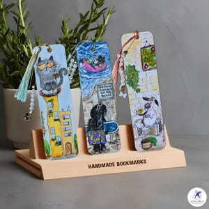 Op de afbeelding: Drie handgemaakte bladwijzers met kleurrijke illustraties van een kat, een hond en een hond op een strand. De bladwijzers staan op een houten standaard met de tekst "HANDGEMAAKTE BLADWIJZERS".