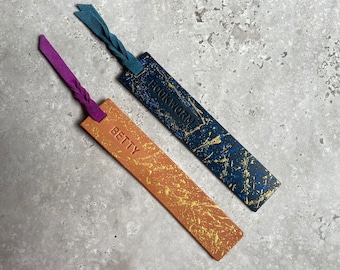 May include: Two leather bookmarks with gold and blue paint splatter designs. One bookmark says "BETTY" and the other says "BOOKWORM".