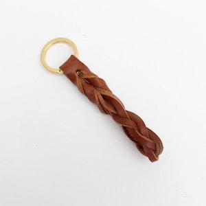 Puede incluir: Un llavero de cuero marrón con un diseño trenzado y un anillo de llave dorado.