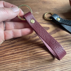 Puede incluir: Un llavero de cuero burdeos con un anillo de llave dorado y un remache de latón. El llavero está sujeto en una mano.