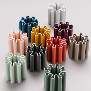 Può includere: Un set di dieci porta matite in plastica colorati a forma di stella. I porta matite sono disposti in cerchio su una superficie grigia. I porta matite sono tutti della stessa dimensione e forma, ma sono di colori diversi, tra cui rosa, giallo, rosso, grigio, verde e viola.