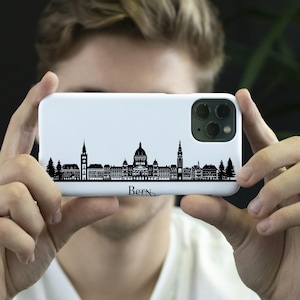 May include: White phone case with a black skyline silhouette of a city, and the word "Bern" at the bottom. The case is held up in front of a person's face, displaying the design. The phone case has a camera bump on the back.