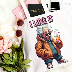 May include: A white tote bag with the words "I LOVE IT" and a cartoon image of Albert Einstein holding a drink. The bag is surrounded by a pink notebook, sunglasses, a smartphone, and pink sneakers.