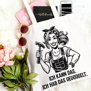 以下が含まれることがあります： ドイツ語のテキストが入った、ハンマーを持った女性の白黒イラストが描かれた白いトートバッグ。バッグの中には、ピンクのノート、黒のノート、スマートフォンが入っています。ピンクのサングラス、ピンクのスニーカー、花も写っています。