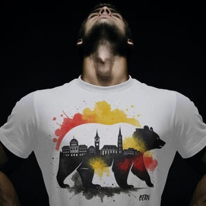 Könnte beinhalten: Weißes T-Shirt mit einer schwarzen Bärensilhouette und einem Stadtbilddesign. Der Bär ist mit einer Stadtlandschaft gefüllt und von roten, gelben und schwarzen Aquarellspritzern umgeben. Das Wort "BERN" ist unten aufgedruckt.