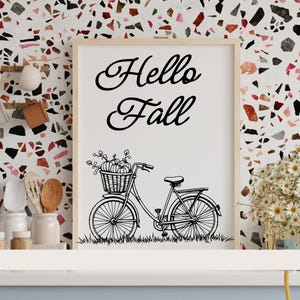 Könnte beinhalten: Ein gerahmter Druck mit dem Schriftzug "Hello Fall" in geschwungener Schrift. Darunter eine schwarz-weiße Illustration eines Fahrrads mit einem Korb voller Kürbisse. Der Rahmen ist hell. Der Hintergrund ist ein farbenfrohes Terrazzo-Muster.