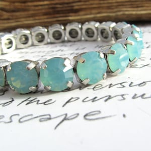 Peut inclure: Un bracelet extensible en argent avec des pierres opalescentes bleu clair. Le bracelet est sur une surface blanche avec du texte manuscrit.