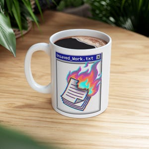 May include: White ceramic coffee mug filled with black coffee. The mug displays a graphic of burning documents with the text "Unsaved_Work.txt" in a retro computer window style. The flames are in shades of orange, pink, and blue.