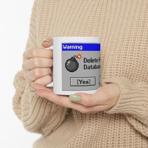 May include: White ceramic mug featuring a retro computer interface design. The mug displays a warning message with a pixelated bomb graphic and the text "Delete Database?" with a "[Yes]" button. The mug is held by a person wearing a beige sweater.