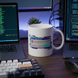 以下が含まれることがあります： 白いセラミックマグカップ、ハンドル付き。コンピューターのエラーメッセージ「Brain.exe - Application Error」が表示されています。メッセージは「深刻なカフェイン欠乏が発生しました。システムがシャットダウンしています。」ボタンには「Abort」、「Retry」、「Pour Coffee」が含まれています。